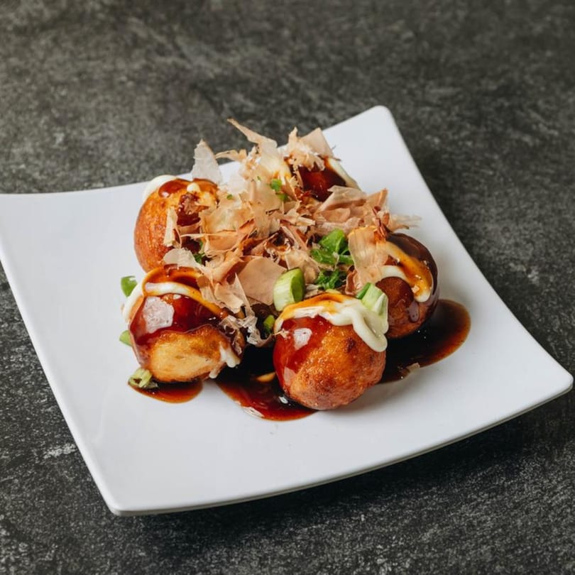 Four golden-brown takoyaki, drizzled with sauce and mayonnaise, topped with bonito flakes and green onions.