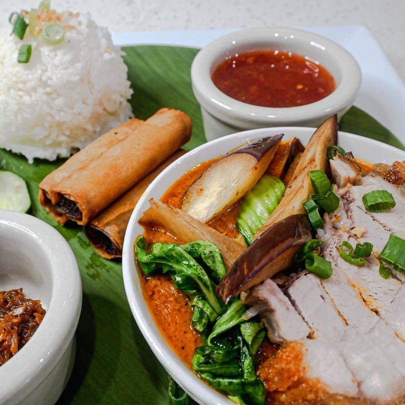 Filipino Kare-Kare with crispy pork belly, vegetables, rice, lumpia, and dipping sauces on a banana leaf.