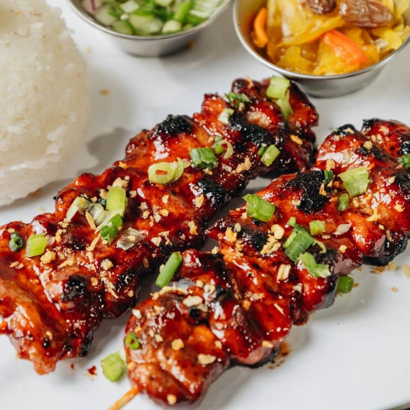 Two skewers of Filipino BBQ pork, garnished with green onions and garlic, served with rice and sides.