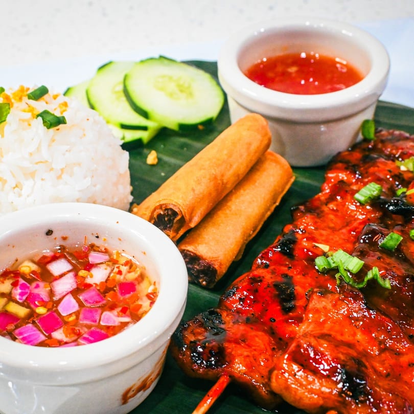 Grilled chicken skewers, two spring rolls, rice, cucumber slices, and two dipping sauces on a banana leaf.
