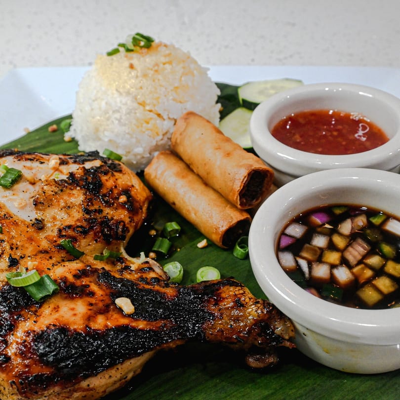Grilled chicken with rice, two spring rolls, cucumber slices, and two dipping sauces on a banana leaf.