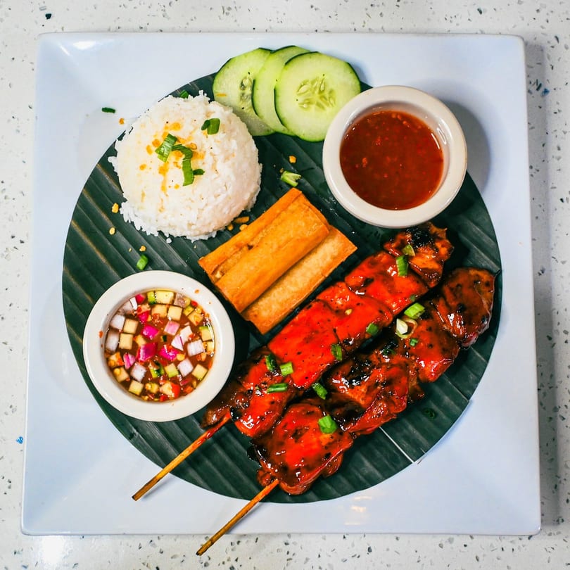 Grilled chicken skewers with rice, spring rolls, cucumber, and two dipping sauces on a banana leaf.