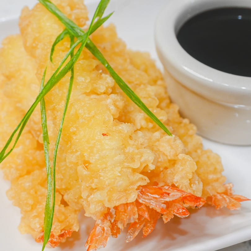 Crispy golden tempura shrimp with green garnish and a small cup of dark dipping sauce.