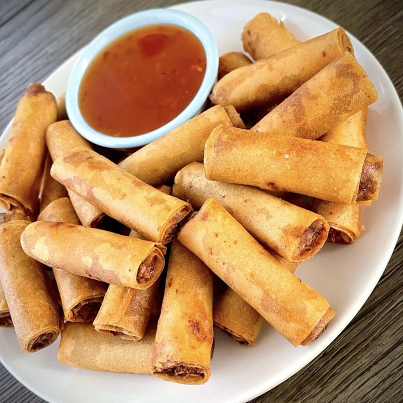 A white plate piled high with golden-brown Filipino lumpia rolls and a small bowl of dipping sauce.