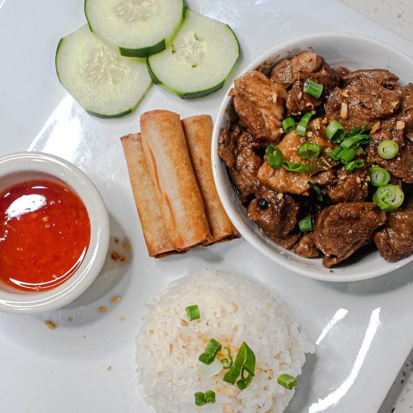 Filipino adobo with rice, lumpia, chili sauce, and cucumber slices on a white plate.