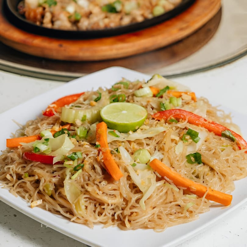 Filipino pancit bihon noodles with vegetables and a lime wedge on a white plate.