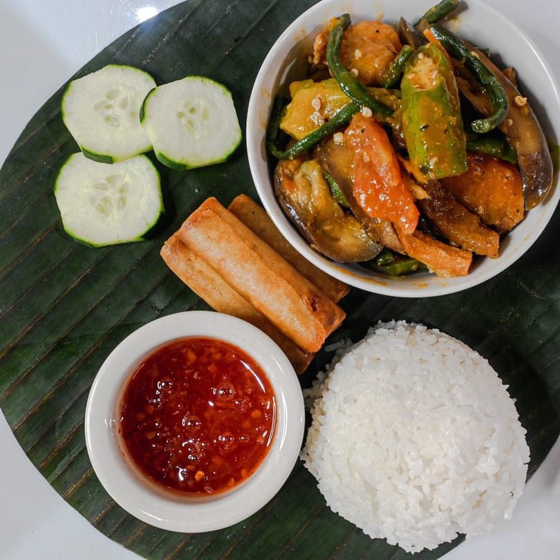Filipino dish Pinakbet with rice, lumpia, fresh cucumbers, and a chili dipping sauce on a banana leaf.