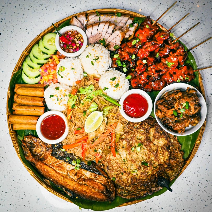 A large platter of Filipino food including grilled fish, skewers, spring rolls, rice, and pancit.